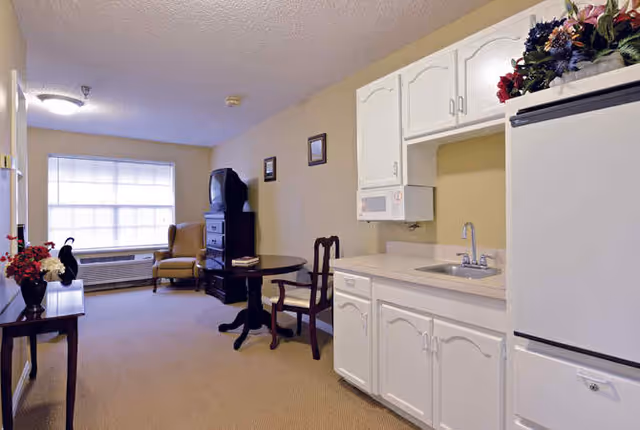 Small studio-style living area with a kitchenette, round table and armchair by a large window.