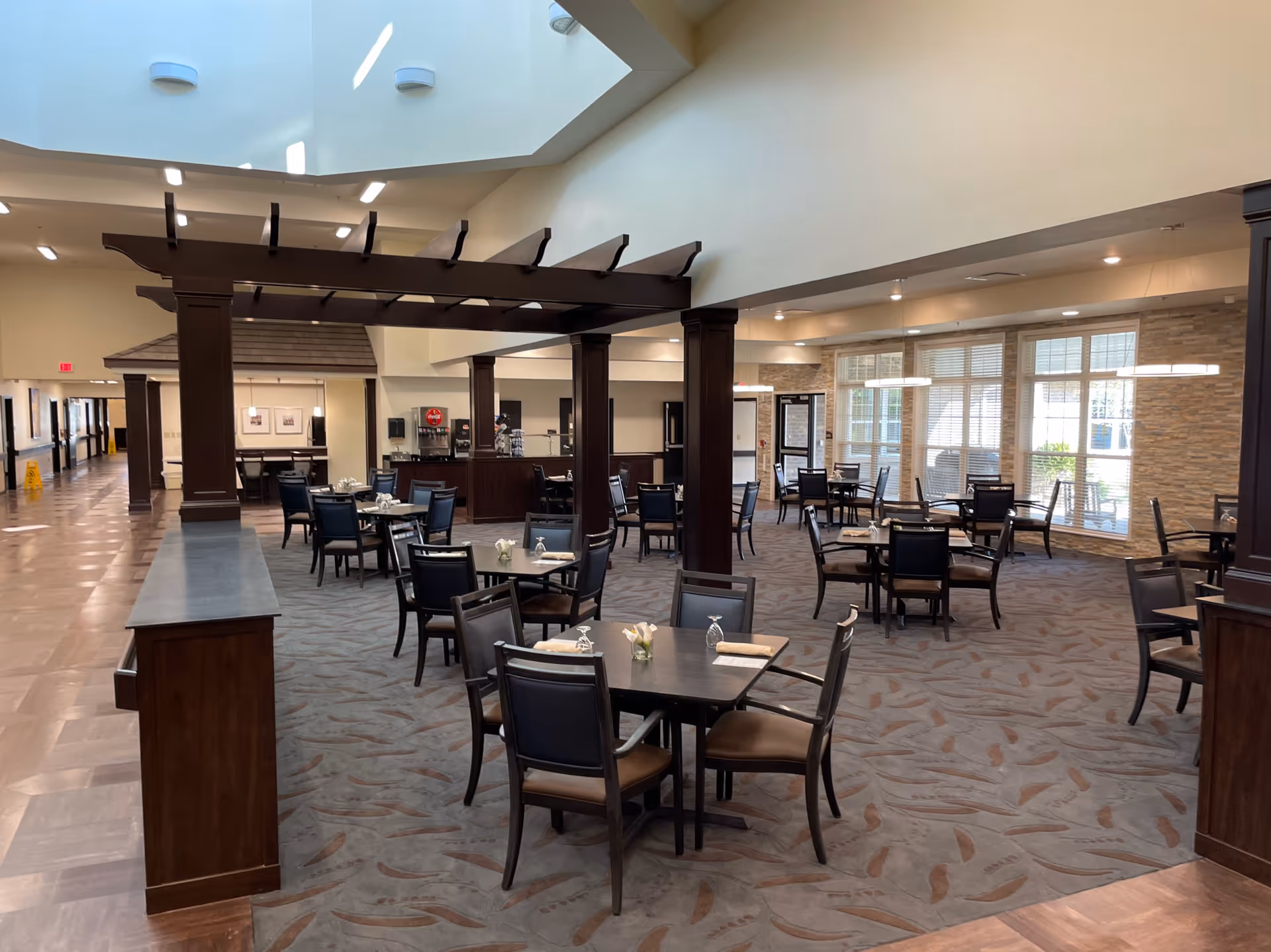 A spacious dining area in a senior living facility with multiple dark wood tables and chairs arranged neatly. The room features a high ceiling with skylights, large windows allowing natural light, and a decorative wooden pergola structure in the center. The floor is carpeted with a patterned design, and there is a beverage station visible in the background.