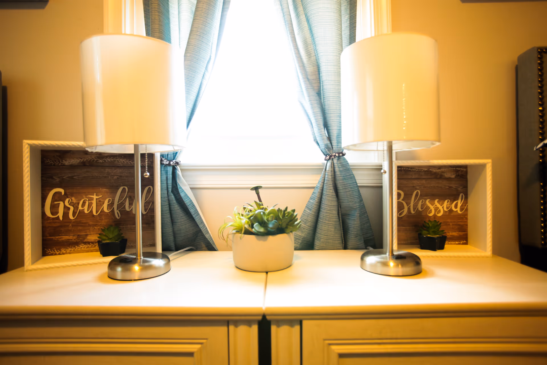 Symmetrical bedside setup with two lamps, draped window, small plant, and grateful/blessed framed signs.