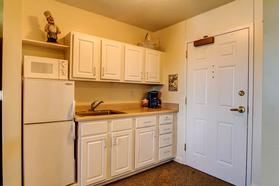 Small kitchen area with white cabinets, a countertop with a sink, a coffee maker, and a refrigerator with a microwave on top. A decorative chef figurine is placed on a shelf above the refrigerator. A white door is visible to the right of the kitchen setup.