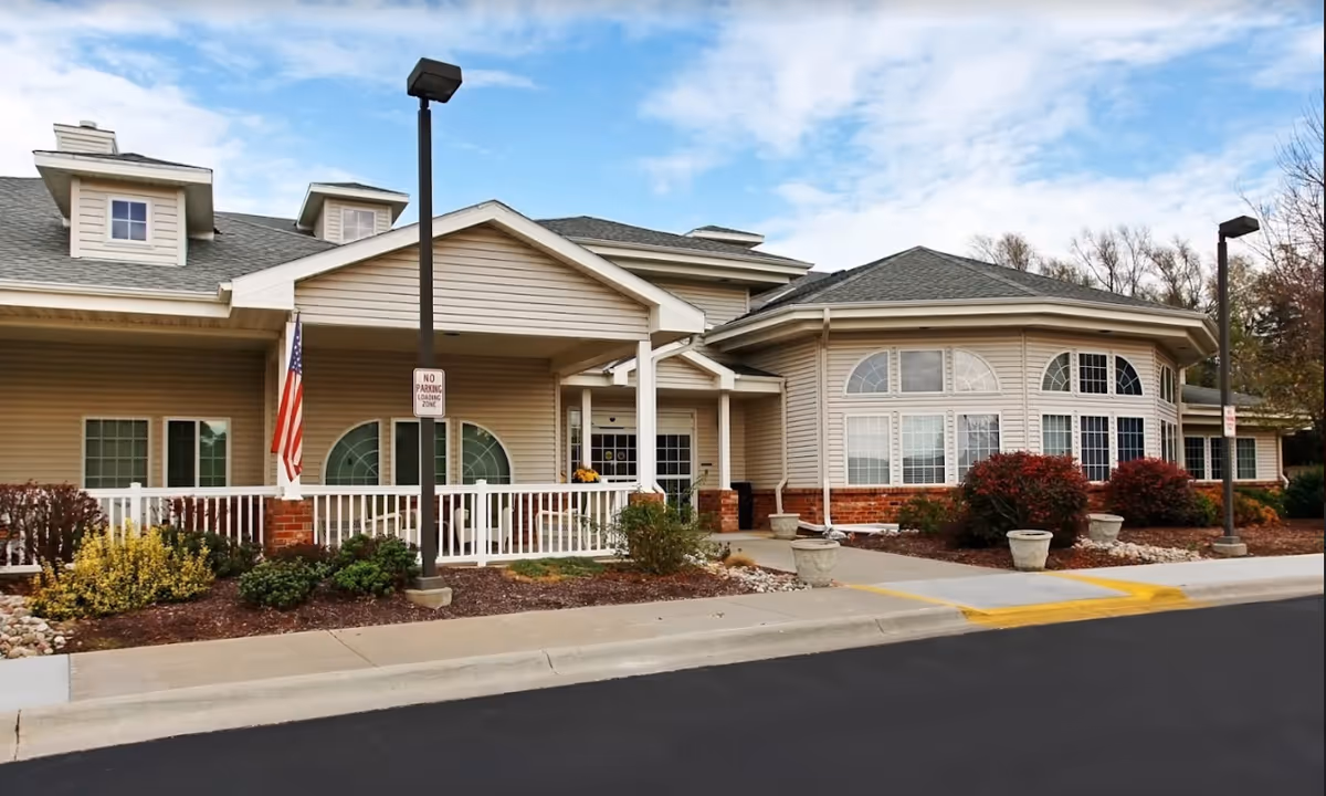 Exterior view of Bailey Pointe Assisted Living at Van Dorn, showing a beige building with white trim, large windows, a covered entrance, landscaped bushes, and a paved driveway with a 'No Parking Loading Zone' sign.
