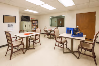 A small activity room with several tables and chairs arranged for group seating. One table has a bingo cage and cards, indicating a recreational activity. The room has a beige floor, white walls, a wooden door, a window looking into another room, a cabinet with a TV on top, and a bookshelf filled with books.