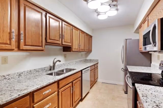 A narrow kitchen with wooden cabinets, granite countertops, a double sink, stainless steel dishwasher, refrigerator, microwave, and electric stove. The floor is tiled and the ceiling has a modern light fixture.