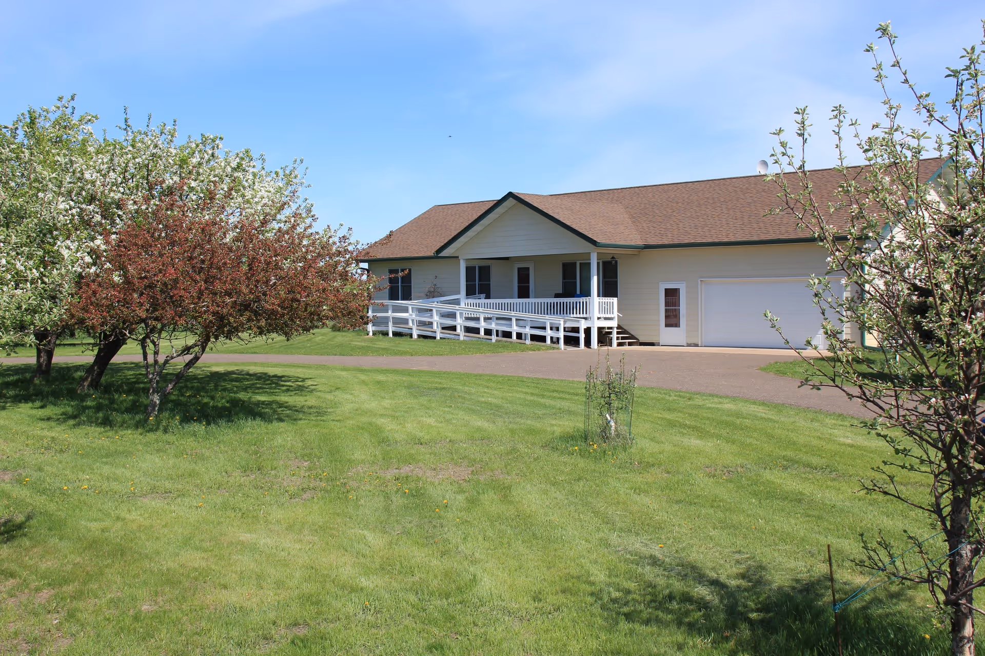 Single-story building with a brown roof and white exterior, featuring a wheelchair accessible ramp and a garage door, surrounded by green grass and blossoming trees under a clear blue sky.