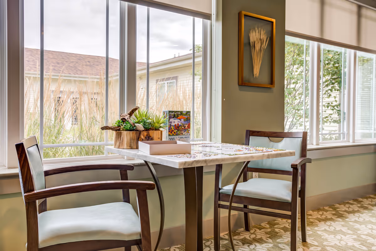 Small seating area with a table and two chairs by large windows, with a puzzle and decorative plants on the table.