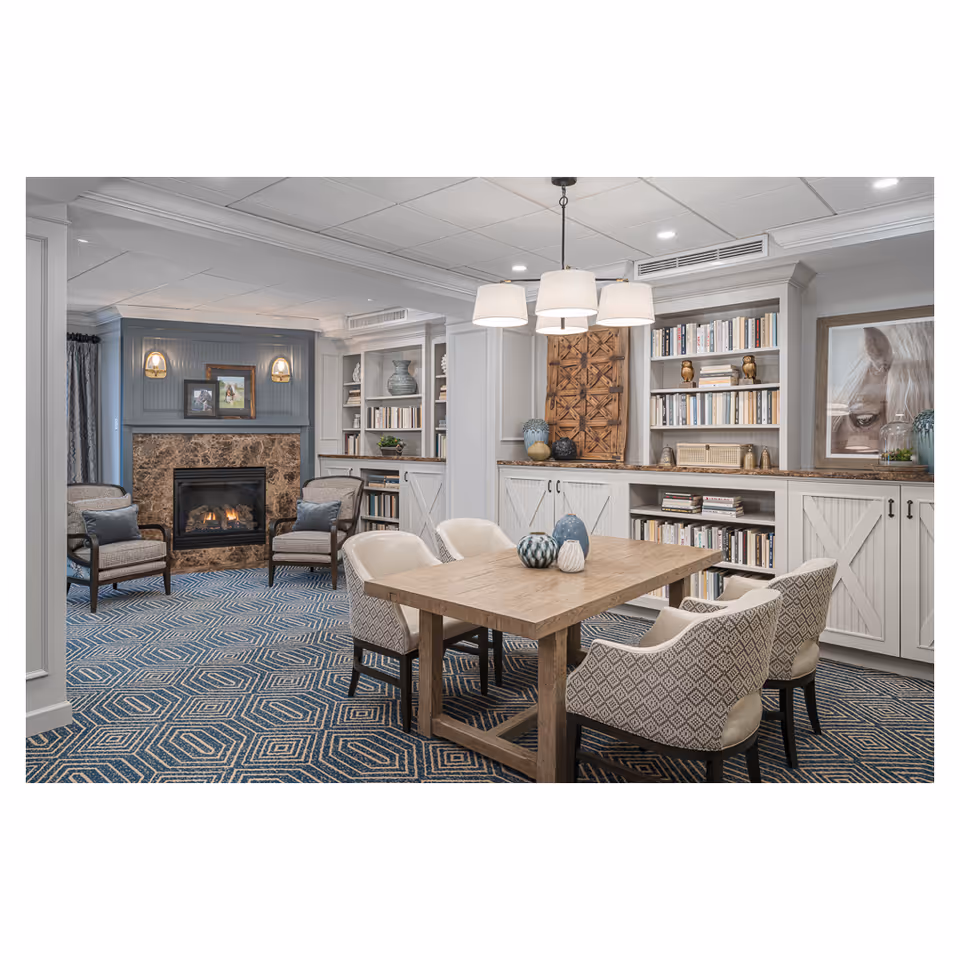 A cozy living room area with a wooden table surrounded by six upholstered chairs. The room features built-in white bookshelves filled with books and decorative items, a fireplace with a marble surround, two armchairs with cushions, and a patterned blue carpet. A modern chandelier with four lamps hangs above the table, and there is a framed picture of a horse on the wall.