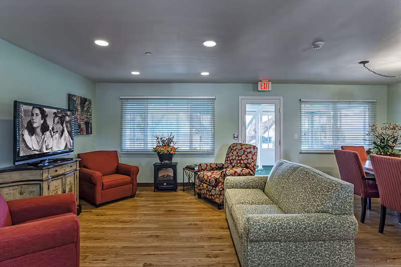 A furnished common room with sofas, armchairs, a television, dining chairs, and windows with an exit door.