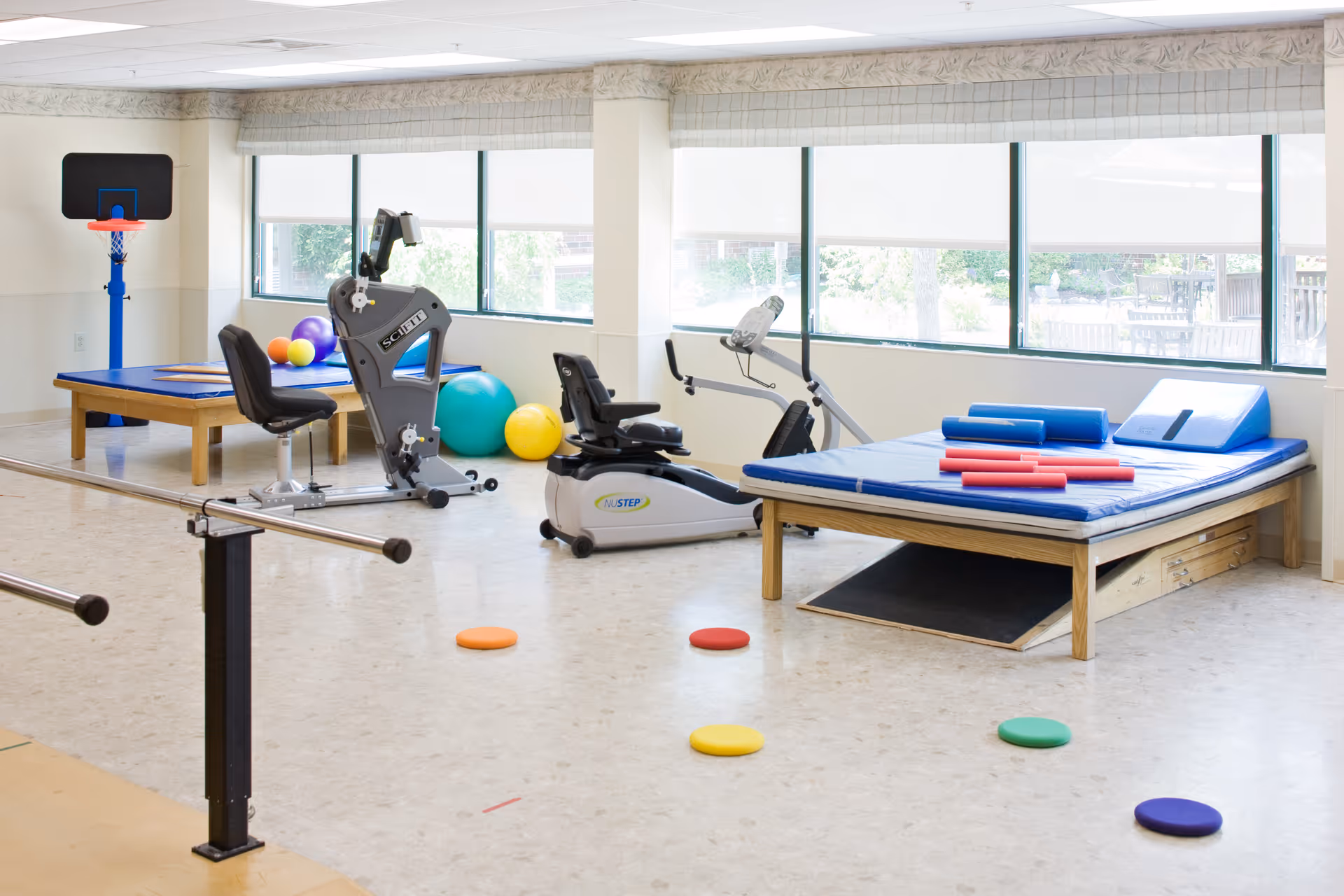 Bright rehabilitation room with exercise bikes, a therapy table and mat, parallel bars, and colorful balance markers.