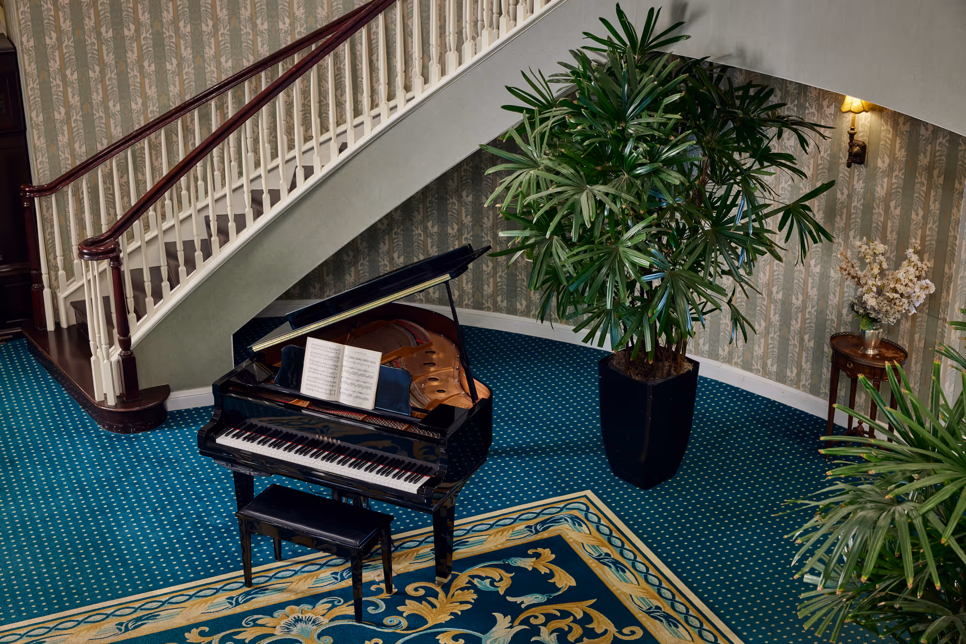 Interior view of a senior living facility featuring a black grand piano with an open music book on the stand, a matching piano bench, a large potted plant, a small round table with a vase of flowers, and a staircase with white railings and dark wooden handrails. The floor is covered with a blue patterned carpet and a decorative rug, and the walls have patterned wallpaper with a wall-mounted light fixture.