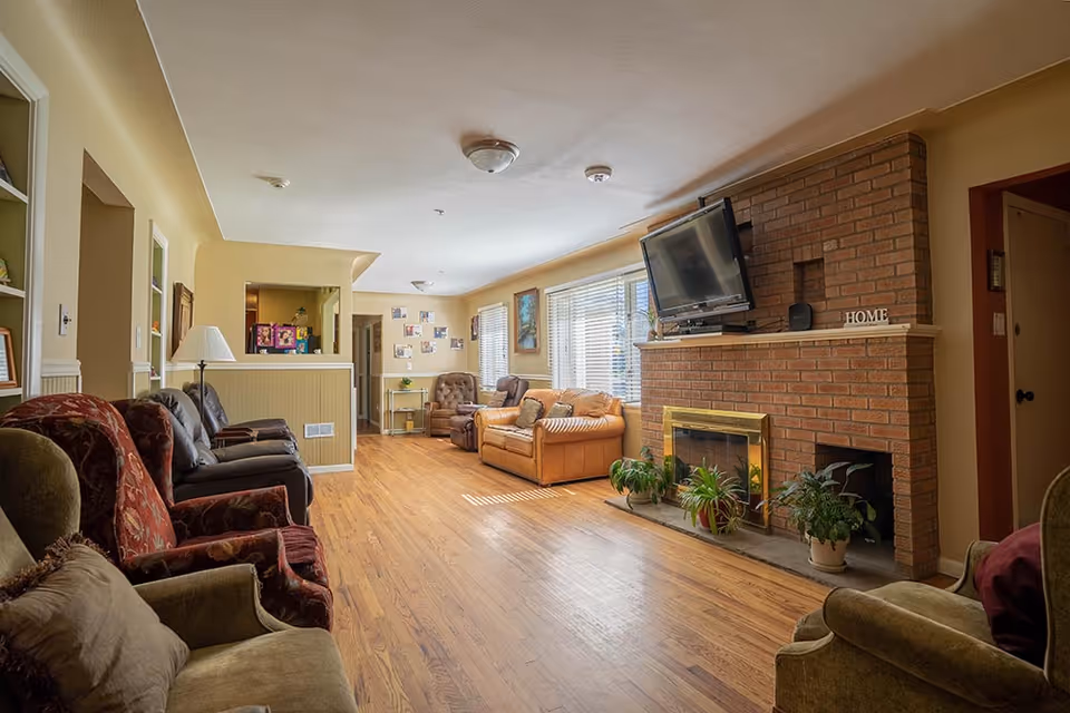 Spacious living room with multiple armchairs and sofas, hardwood floors, a brick fireplace with a mounted TV, and potted plants.