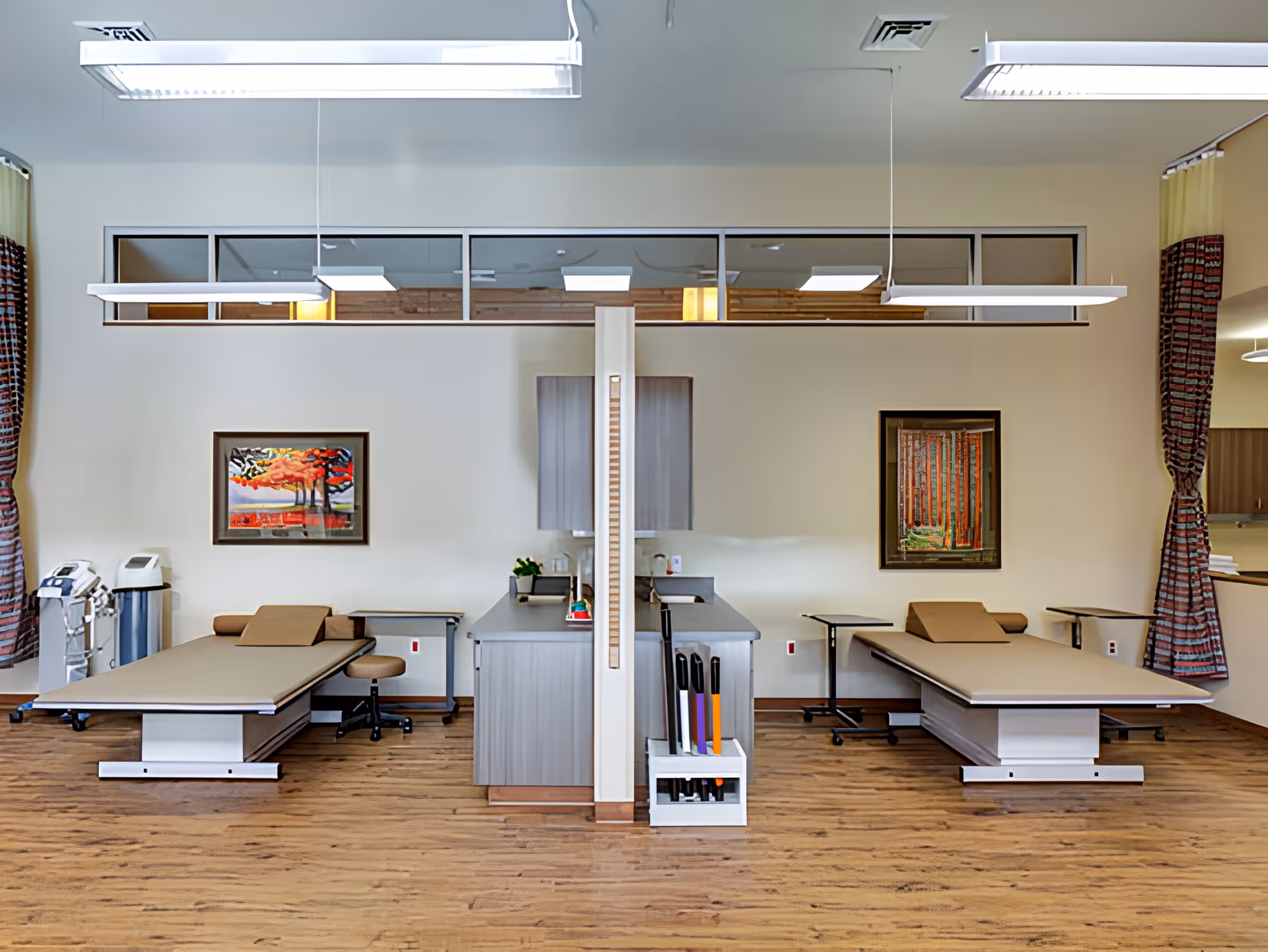 A clean and organized medical treatment room with two examination tables separated by a central cabinet. Each table has a brown cushioned surface and is accompanied by a small stool and a side table. The room features wood flooring, light-colored walls, two framed paintings, and large windows with patterned curtains. Overhead fluorescent lights illuminate the space.