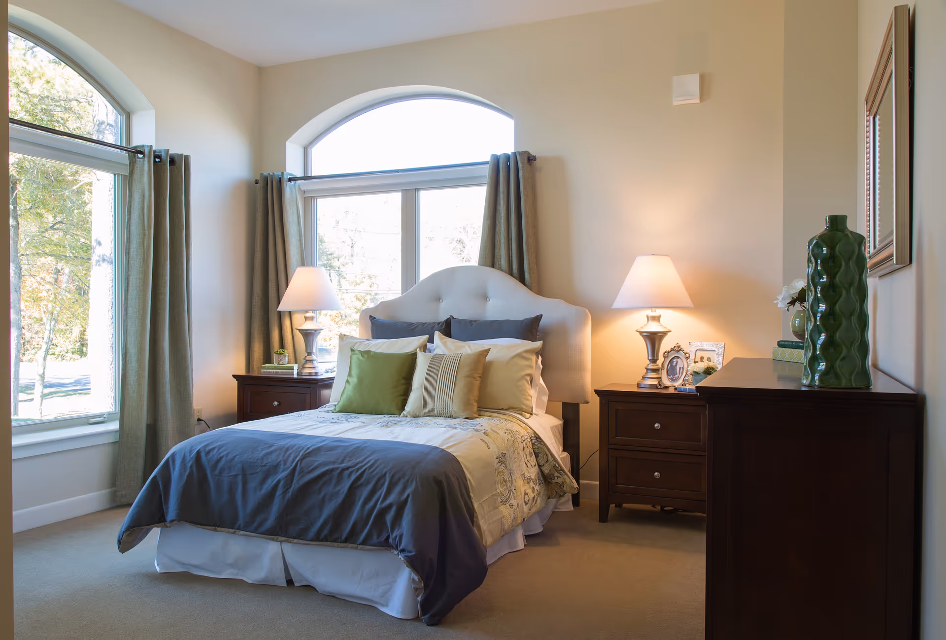 A cozy bedroom with a bed centered under a large arched window. The bed has a tufted headboard and is made with multiple pillows and a comforter in shades of blue, green, and beige. There are two wooden nightstands on either side of the bed, each with a lamp. A dresser with decorative items, including a green vase and framed photos, is on the right side of the room. The room has beige walls and carpet, and large windows with green curtains letting in natural light.