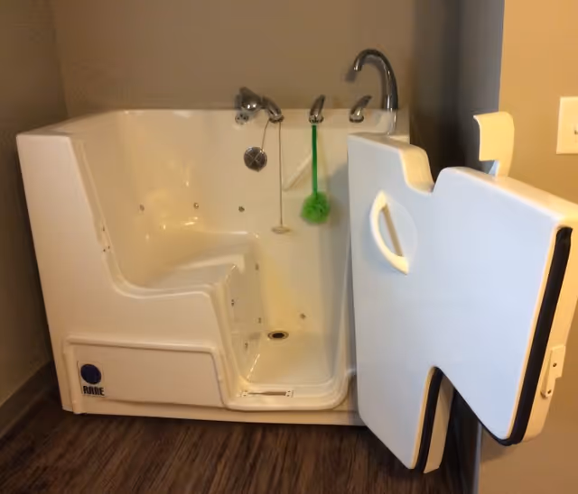 A white walk-in bathtub with its hinged door open, built-in seat, and faucet against a gray wall.