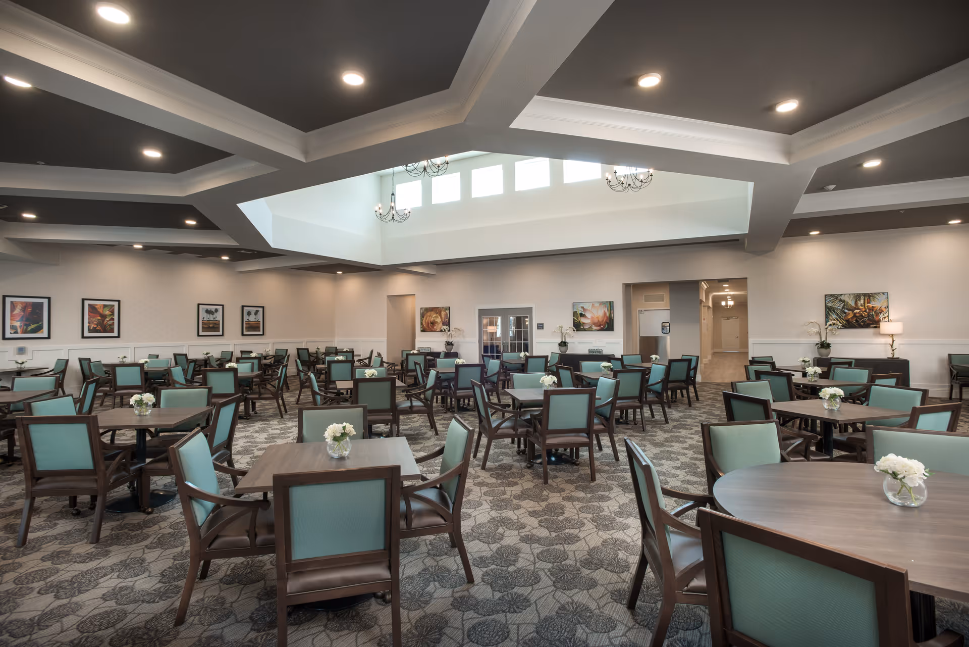 A spacious dining room with multiple wooden tables and teal cushioned chairs arranged neatly. The room has a patterned carpet, framed artwork on the walls, and a large skylight ceiling with chandeliers providing natural light. Small flower arrangements are placed on each table.