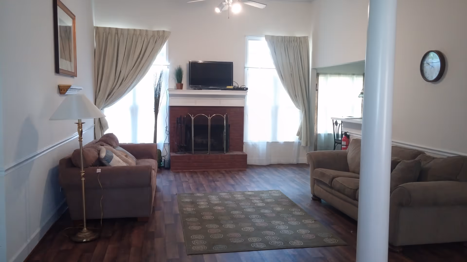 A living room with two brown sofas facing each other, a floor lamp beside one sofa, a fireplace with a TV on the mantel, two tall windows with curtains on either side of the fireplace, a green rug with circular patterns on the wooden floor, and a wall clock on the right wall.