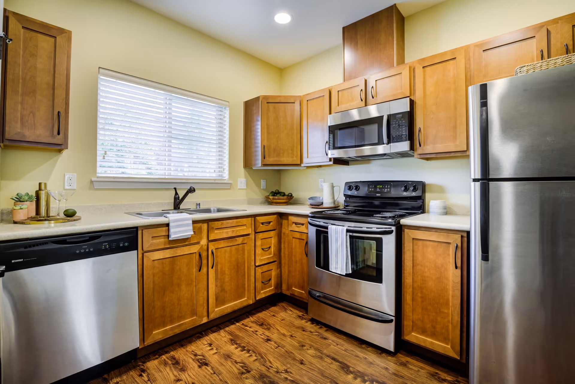 A modern kitchen with wooden cabinets, stainless steel appliances including a refrigerator, microwave, stove, and dishwasher. There is a window with blinds above the sink, and the floor is made of dark wood.