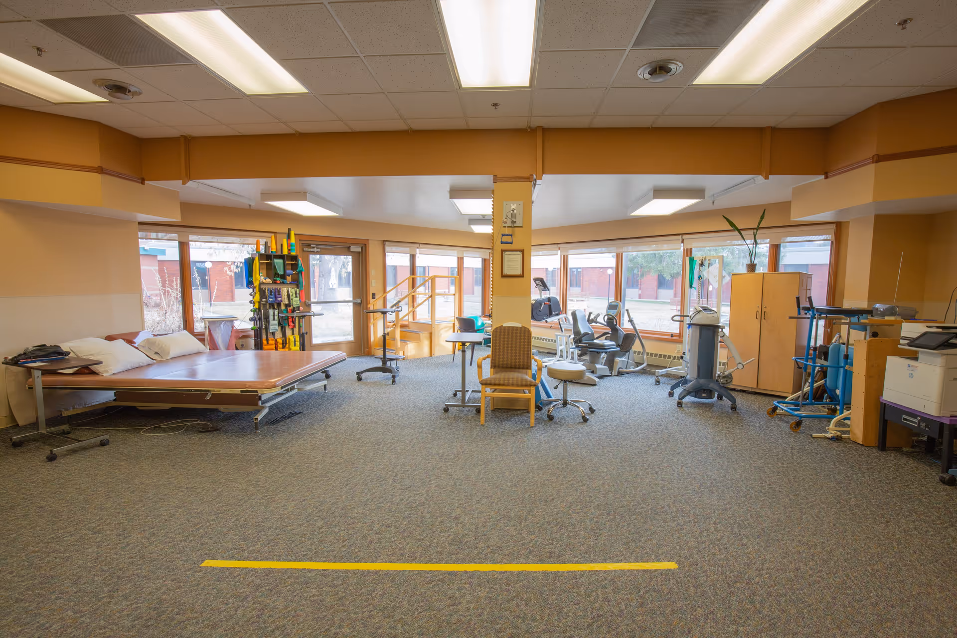 A spacious room with large windows letting in natural light, featuring a hospital bed with pillows on the left, various medical and therapy equipment including exercise machines and chairs, a cabinet with a plant on top, and a carpeted floor with a yellow line marking.