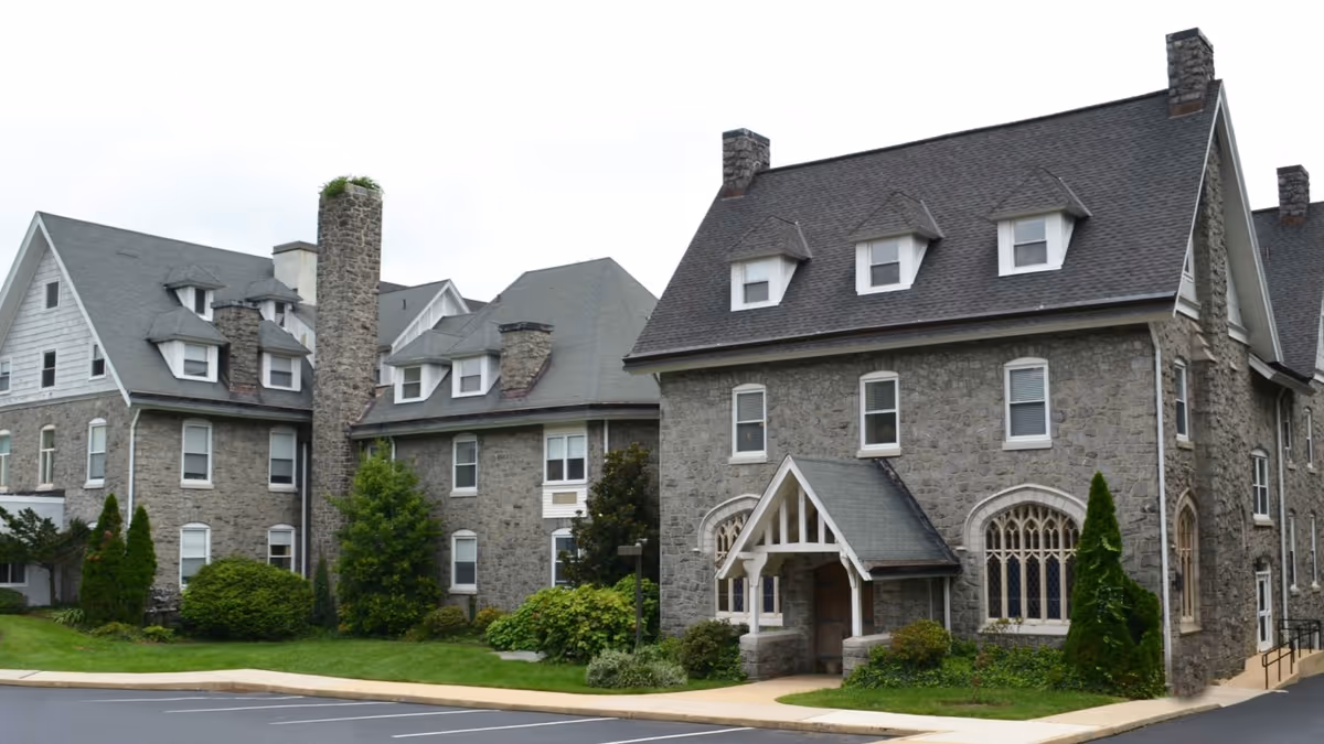 Exterior view of a large stone building with multiple chimneys and dormer windows, surrounded by green shrubs and trees, with a paved driveway in front.