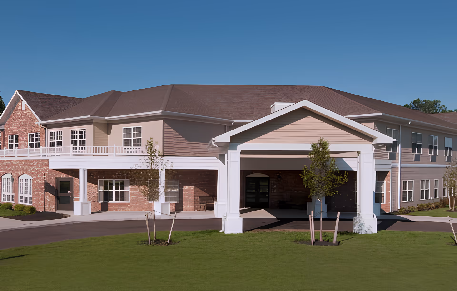 Front entrance of a two-story assisted living building with a covered porte-cochère, brick and siding facade, and a landscaped lawn.