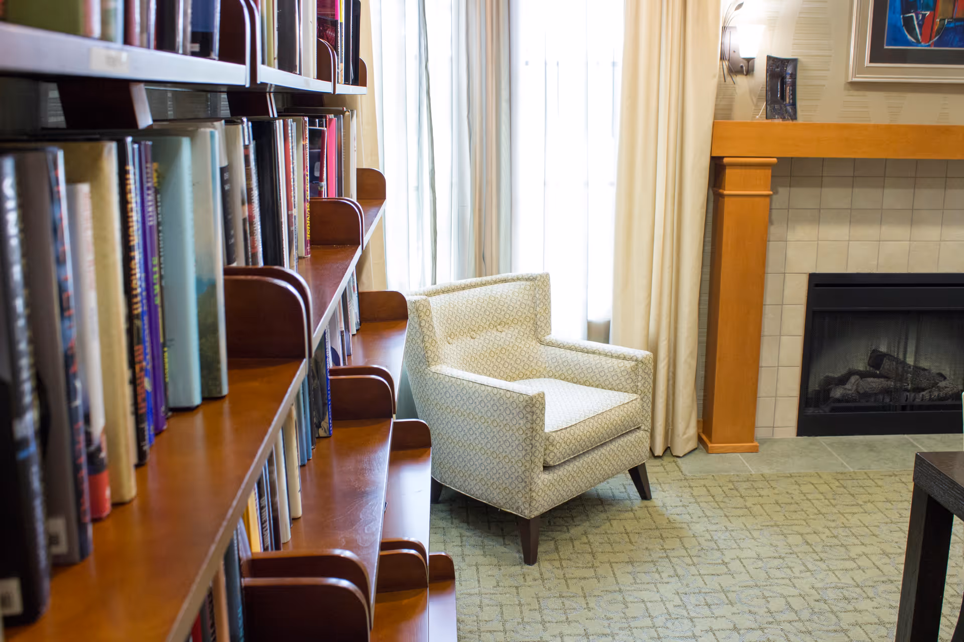 A patterned armchair sits beside a bookshelf and a fireplace in a carpeted lounge area with curtains.