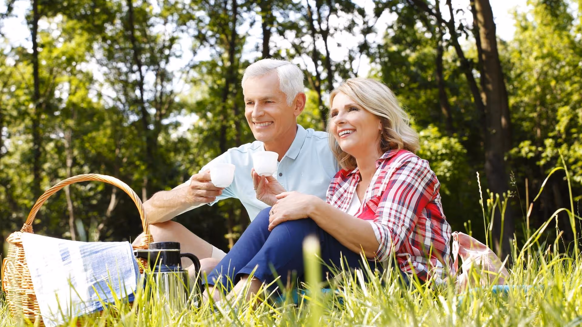 An elderly couple sitting on the grass in a park having a picnic. They are smiling and holding white cups, with a picnic basket and a thermos nearby. The background shows green trees and sunlight filtering through.