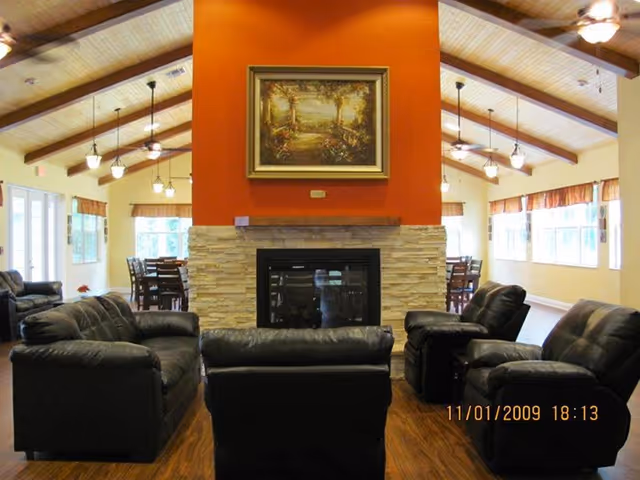 Common room with black leather sofas arranged around a central stone fireplace beneath a vaulted ceiling with exposed beams and dining tables in the background.