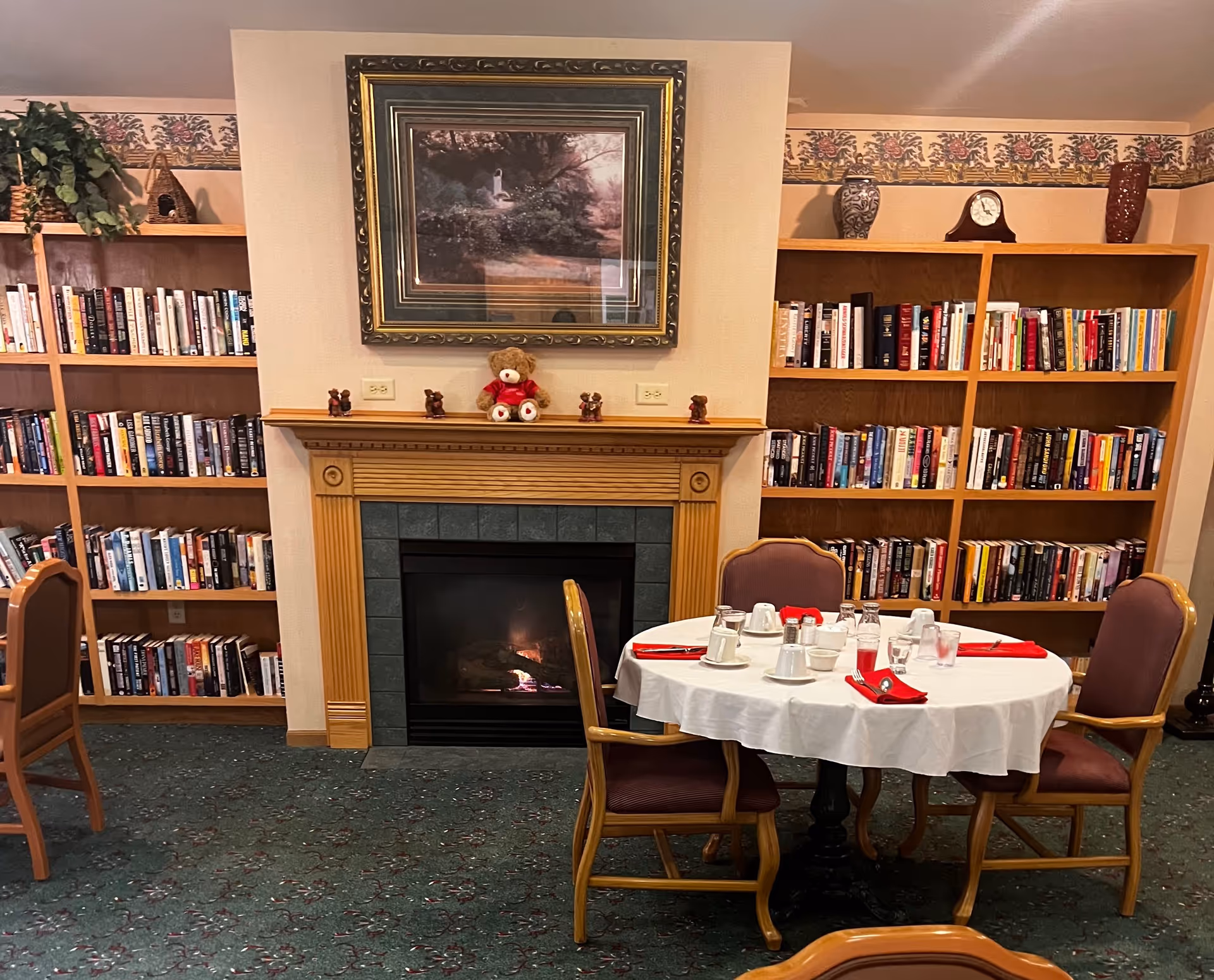 A cozy room with a lit fireplace in the center, flanked by two wooden bookshelves filled with books. Above the fireplace is a framed painting and a teddy bear with smaller bear figurines on the mantel. In front of the fireplace is a round table set with white tablecloth, cups, glasses, and red napkins, surrounded by four wooden chairs with cushioned seats.