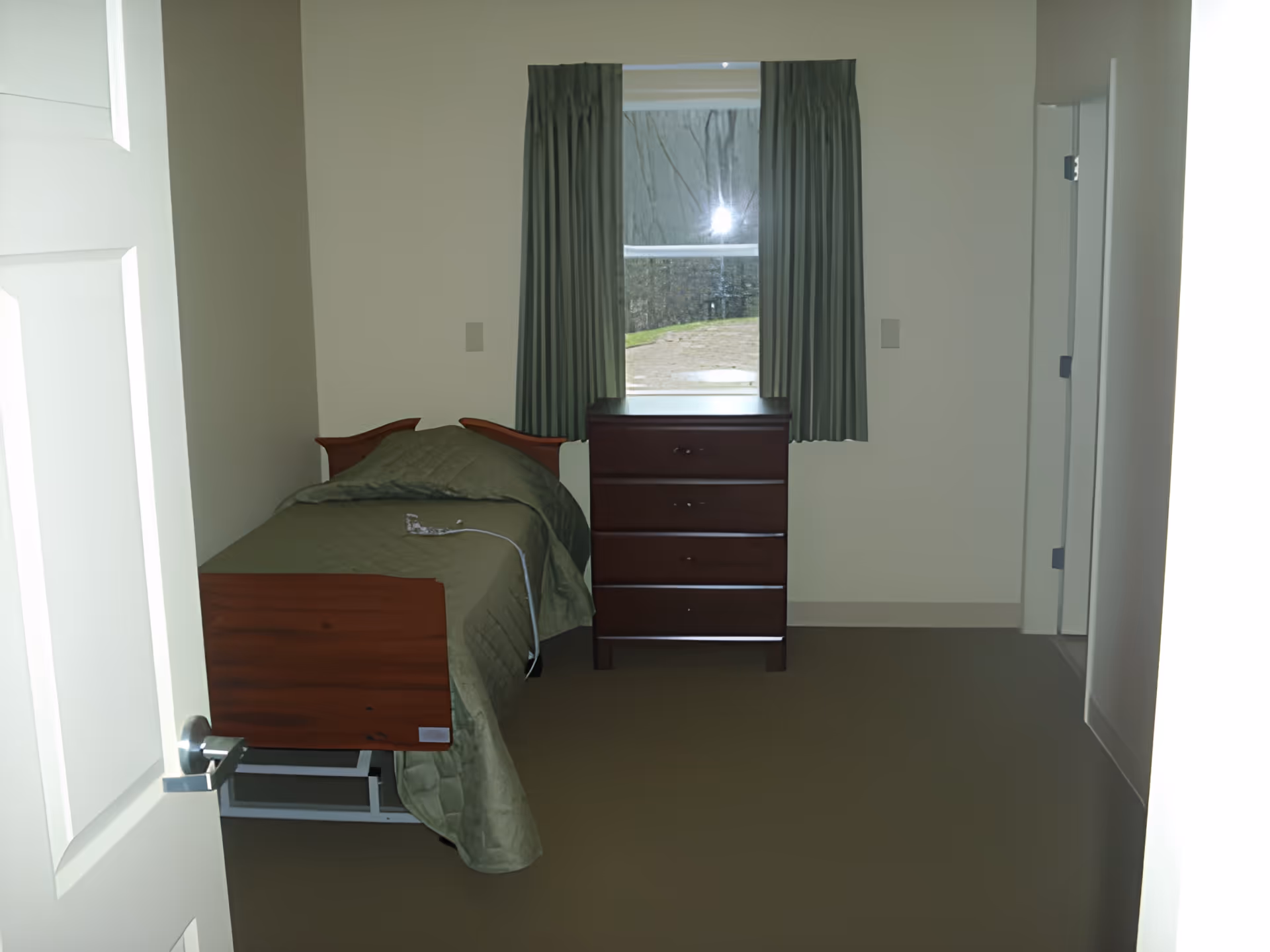 A small bedroom with a single bed covered in a green blanket, a wooden dresser with three drawers, and a window with green curtains. The room has beige walls and a brown floor, with a door partially open on the left side.