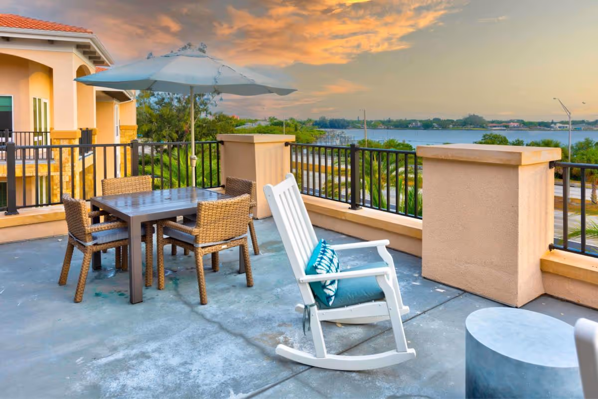 Outdoor patio area with a table and four wicker chairs under a large umbrella, a white rocking chair with a blue cushion, and a view of greenery and water in the background during sunset.
