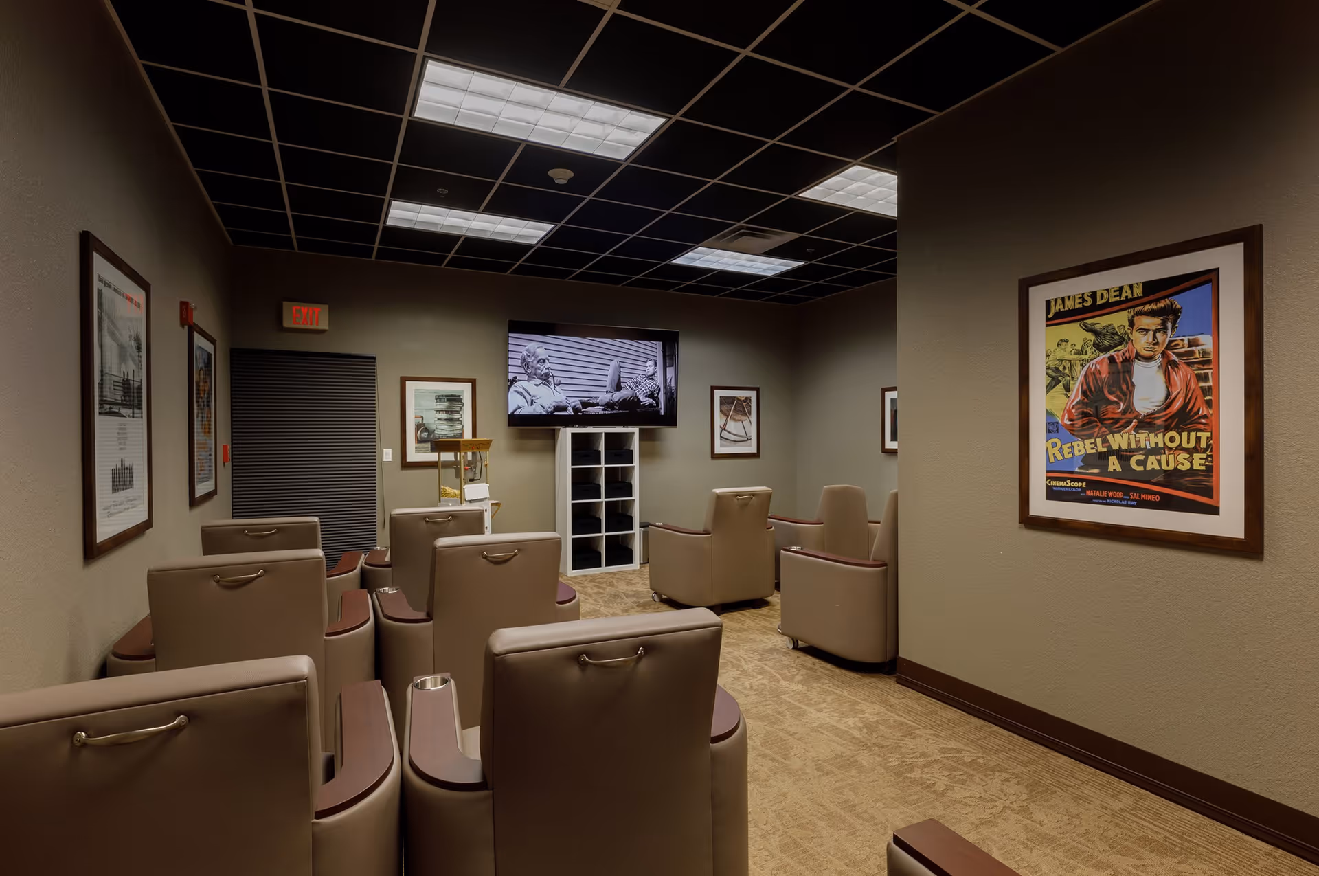 A small theater room with several beige recliner chairs arranged in rows facing a wall-mounted flat screen TV displaying a black and white movie scene. The room has a dark grid ceiling with fluorescent lights, beige walls adorned with framed vintage movie posters, including one of James Dean's Rebel Without a Cause, and a carpeted floor.