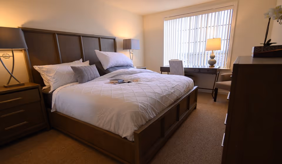 A cozy bedroom with a large wooden bed featuring white bedding and multiple pillows. There are two bedside tables with lamps on each side of the bed. A desk with a chair and a lamp is positioned in front of a large window with vertical blinds, and a dresser is visible on the right side of the room.