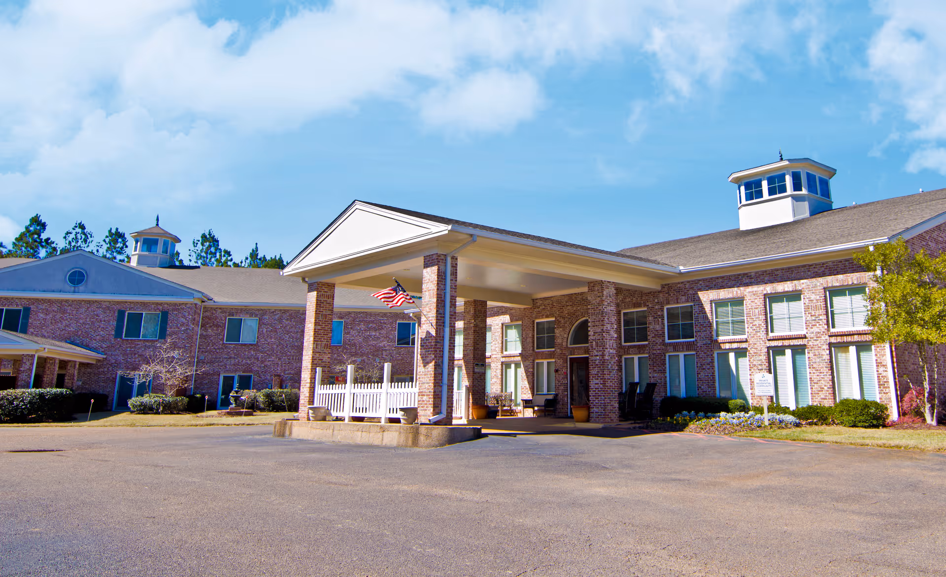 Exterior view of a large brick senior living facility building with a covered entrance supported by brick columns, an American flag hanging, and a clear blue sky above.
