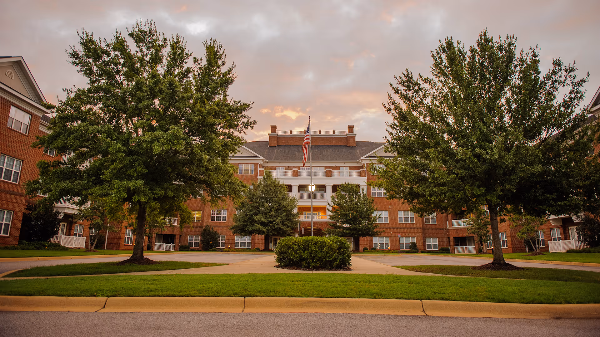 Front exterior view of a large brick building with multiple windows and white balconies, surrounded by green trees and a well-maintained lawn under a cloudy sky during sunset. An American flag is displayed on a flagpole in front of the building.