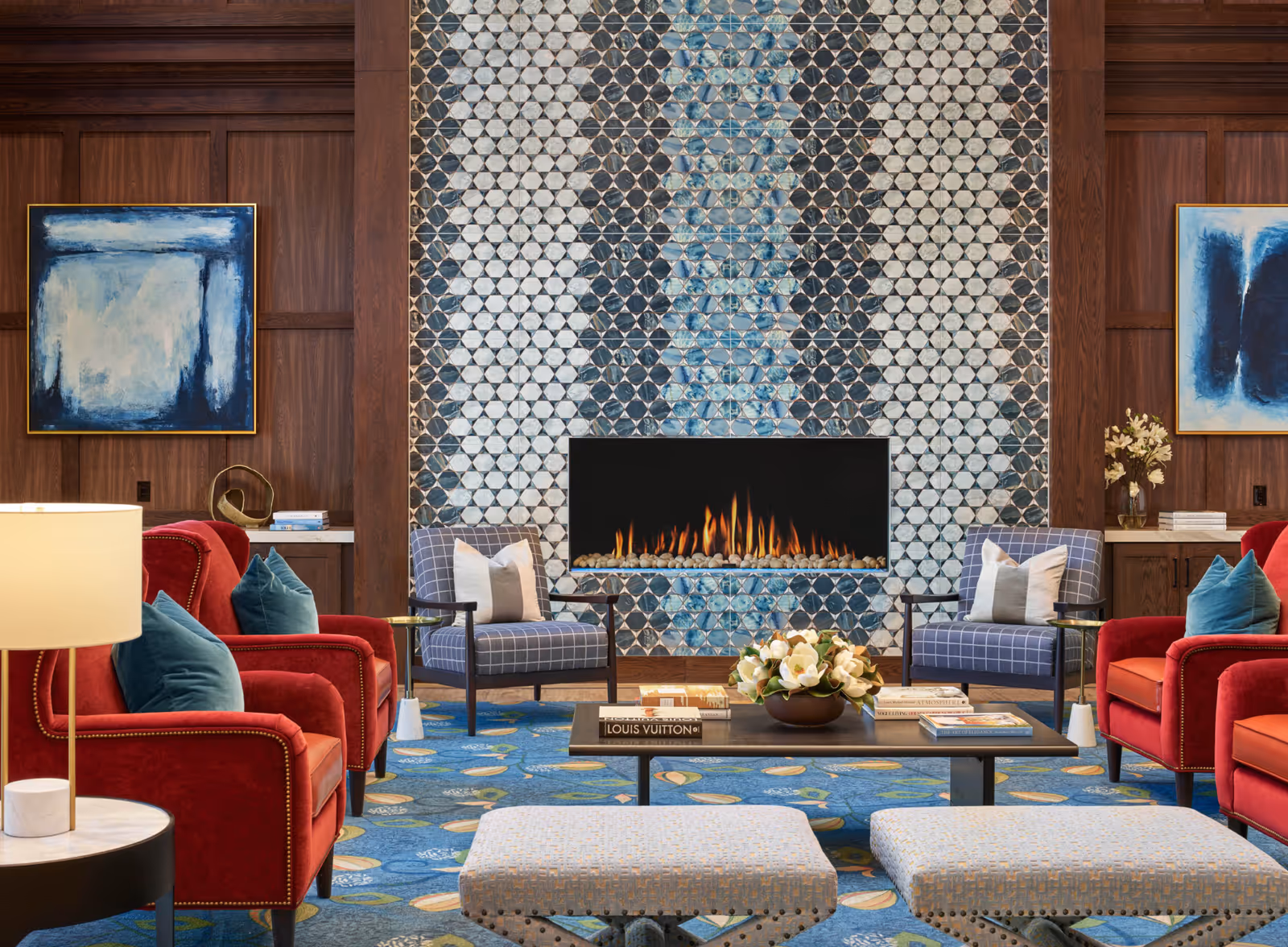 Cozy seating area with a decorative tiled fireplace flanked by wood paneling, red armchairs and blue accent chairs around a coffee table with books and a floral arrangement.