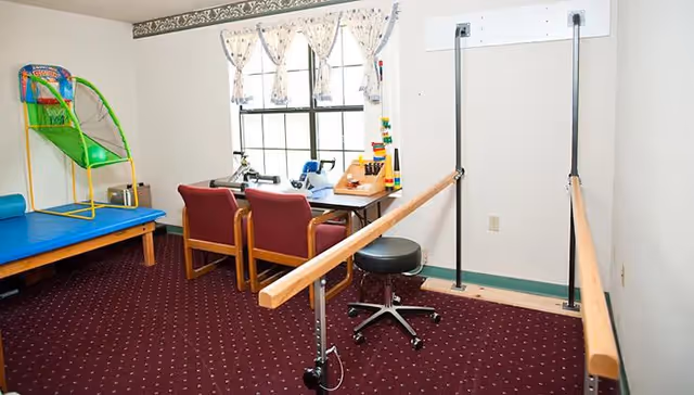 A therapy or activity room with a small basketball hoop game, a padded blue table, two red chairs at a table with various therapy or activity tools, and parallel wooden bars for walking exercises. The room has a window with floral curtains and a maroon carpet with a dotted pattern.