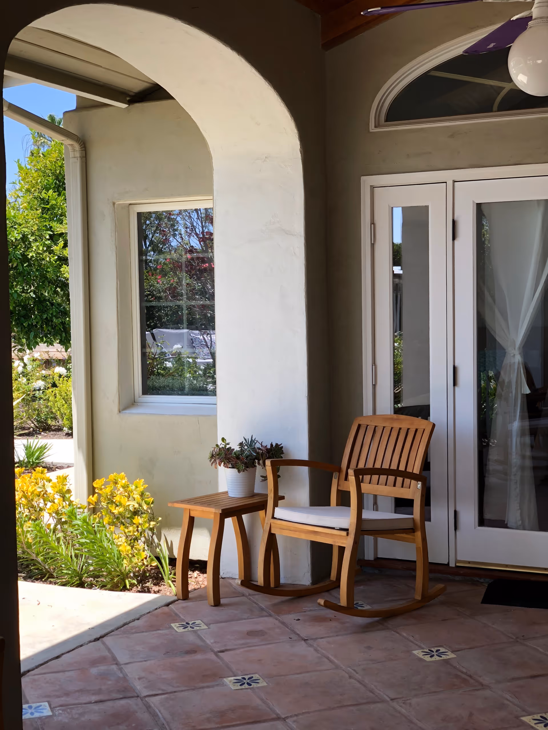 A covered outdoor patio area with a wooden rocking chair and a small wooden side table holding a potted plant. The patio has tiled flooring with decorative tiles, an arched wall opening, a window, and a set of white French doors with glass panels and curtains inside. There are green plants and yellow flowers visible outside in the garden area.