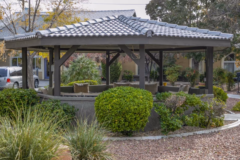 Outdoor gazebo with a tiled roof and cushioned seating inside, surrounded by green bushes and plants, with a building and parked cars visible in the background.