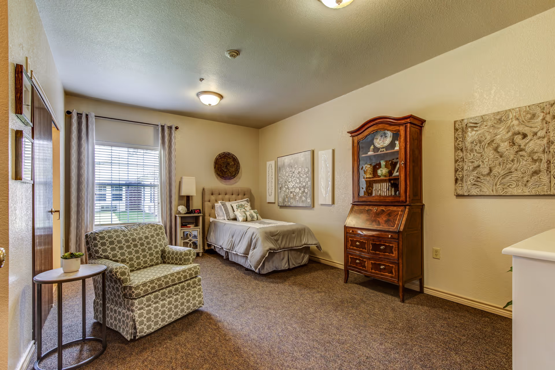 A senior living bedroom featuring a single bed with gray bedding, a patterned armchair next to a small round table with a potted plant, a wooden cabinet with glass doors displaying decorative items, and wall art. The room has a window with blinds and curtains, beige walls, and carpeted flooring.