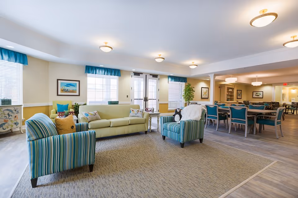 A bright and spacious common area in a senior living facility featuring a seating area with a light green sofa and two striped armchairs with colorful pillows. Behind the seating area, there is a dining space with multiple tables and teal chairs. The room has large windows with blue valances, light-colored walls, and ceiling lights providing warm illumination.