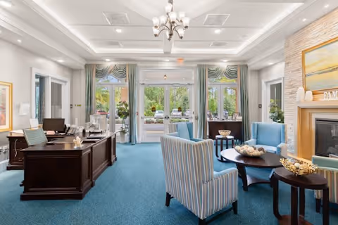 A bright and welcoming interior space with blue carpet and light-colored walls. The room features a dark wooden reception desk on the left with office equipment, and a seating area on the right with striped and solid blue upholstered chairs around dark wooden coffee tables. Large windows and glass doors at the back allow natural light to enter, showing greenery and parked cars outside. The ceiling has recessed lighting and a chandelier, and there is a fireplace with a painting above it on the right wall.
