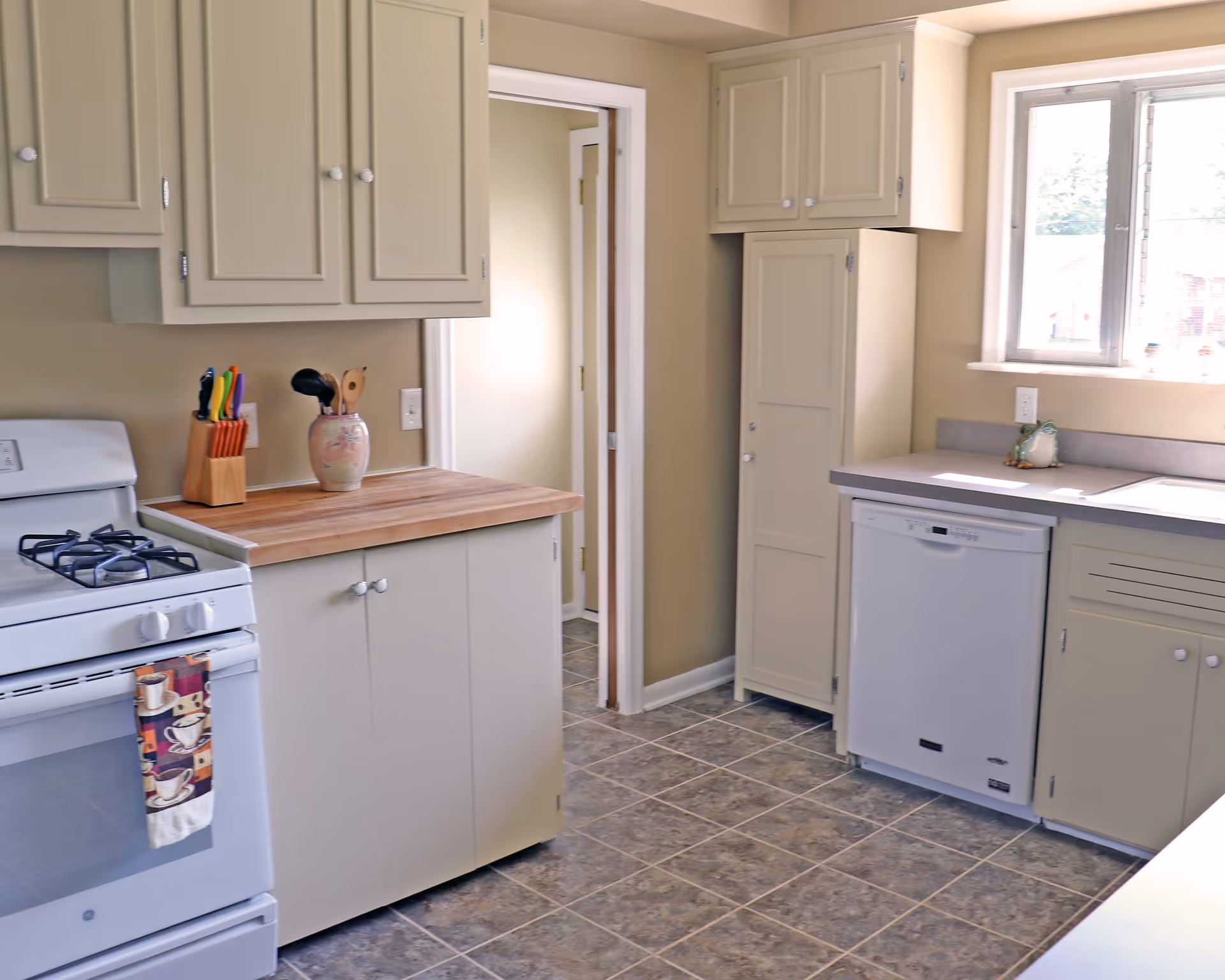 A clean kitchen with beige cabinets, a white gas stove with a coffee-themed towel hanging on the handle, a wooden countertop with a knife block and utensil holder, a dishwasher, and a window letting in natural light.