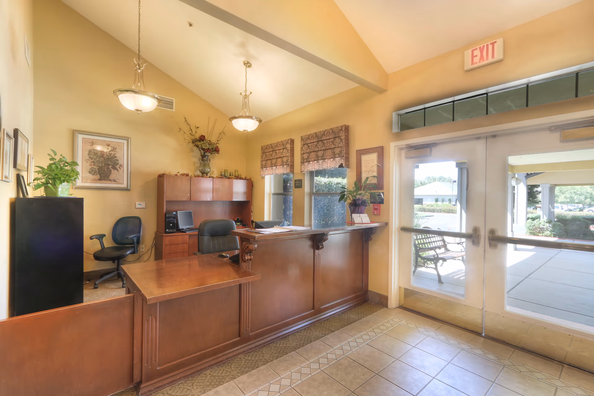 Reception area with a wooden desk and office chairs, decorated with plants and framed artwork on the walls. Two hanging light fixtures illuminate the space. There are windows with patterned valances and a glass exit door leading outside, where a bench and greenery are visible.