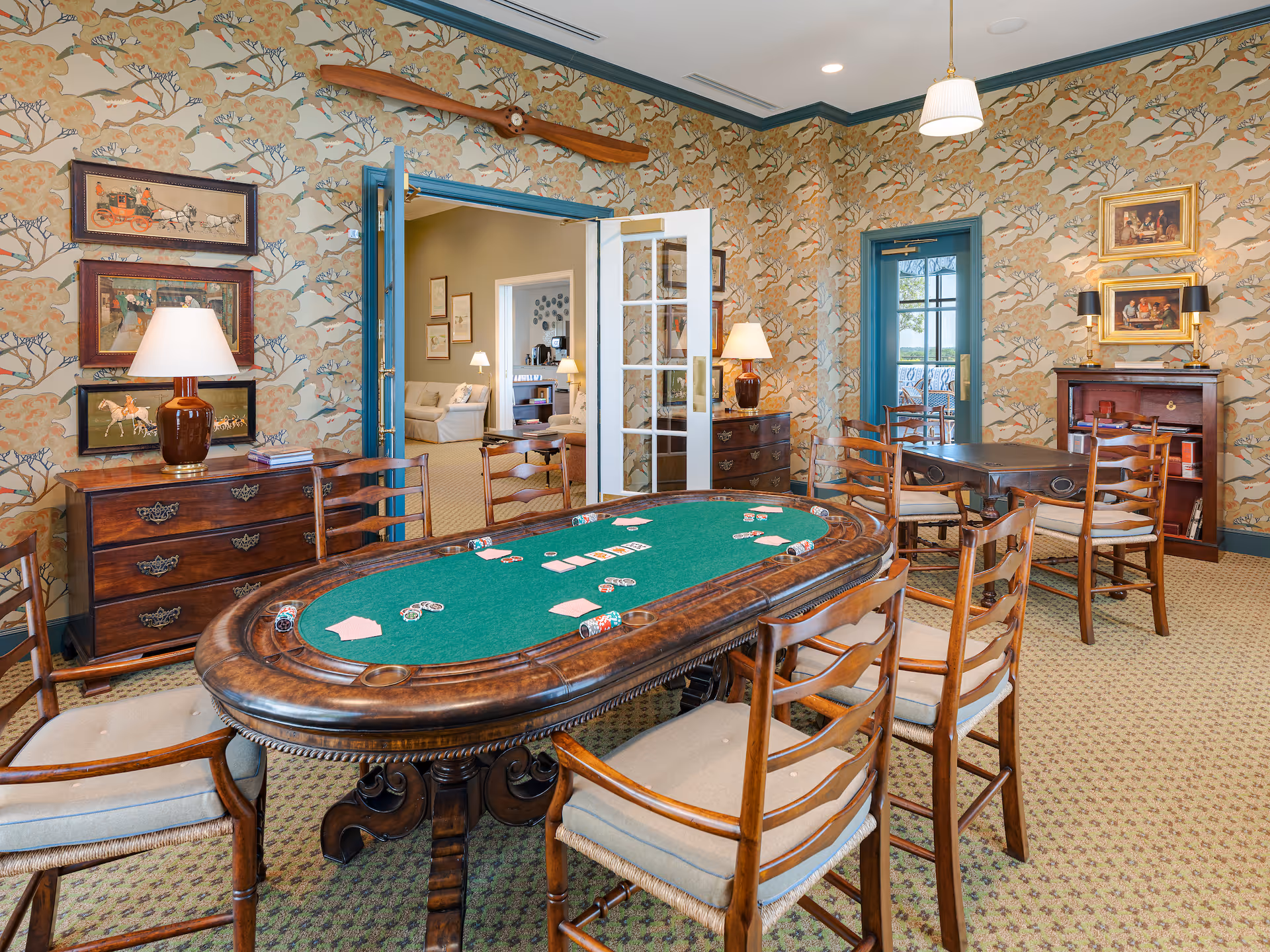 A well-decorated game room with a green felt poker table surrounded by wooden chairs with cushions. The room features floral wallpaper with birds, framed artwork on the walls, two wooden dressers with table lamps, and a wooden propeller mounted above the doorway. French doors lead to an adjacent room with sofas and lamps.