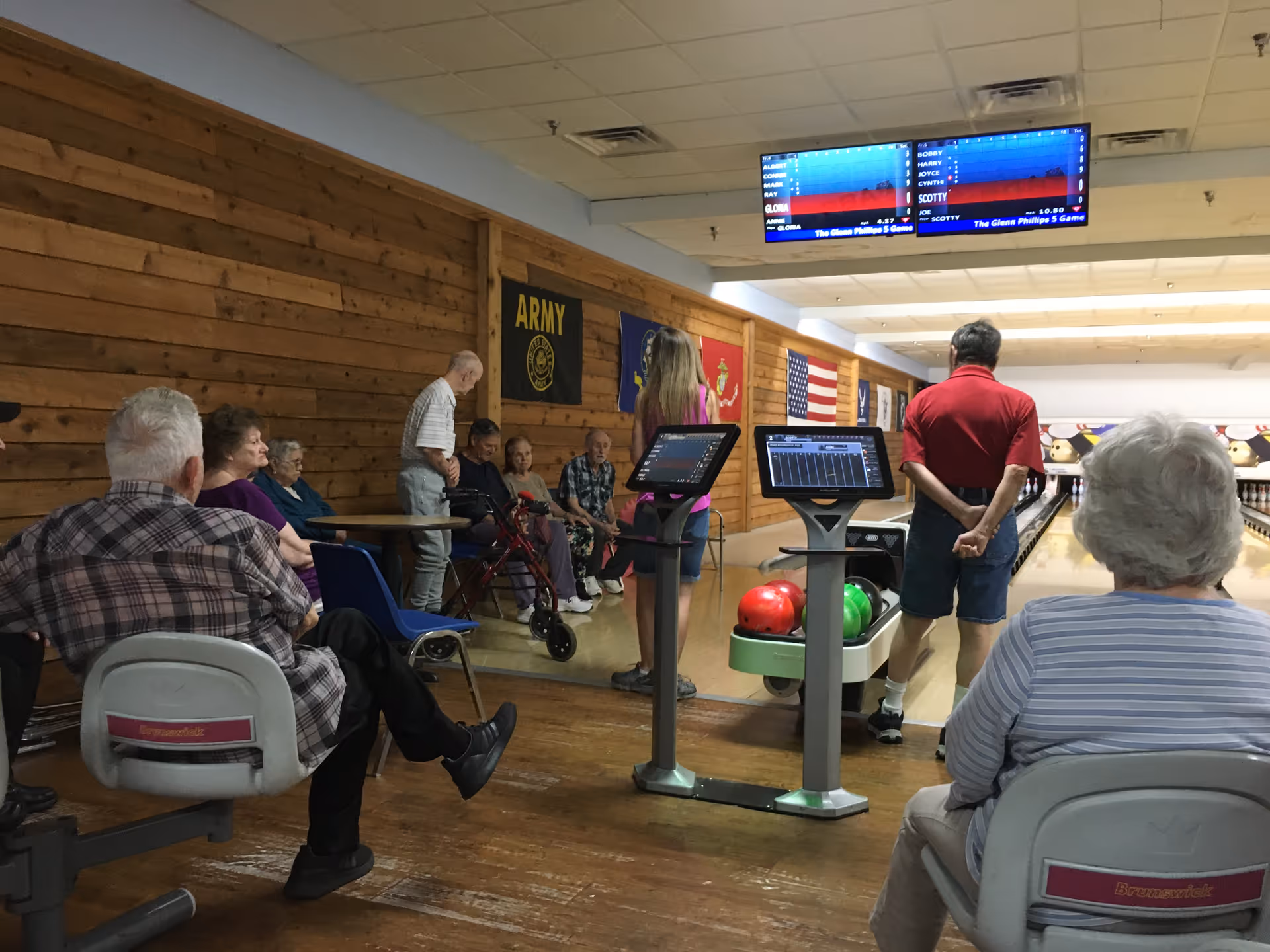 A group of elderly people gathered in a bowling alley. Some are seated on chairs, while others stand near the bowling lanes. The walls are decorated with military flags and an American flag. Two digital scoring screens are visible above the lanes.