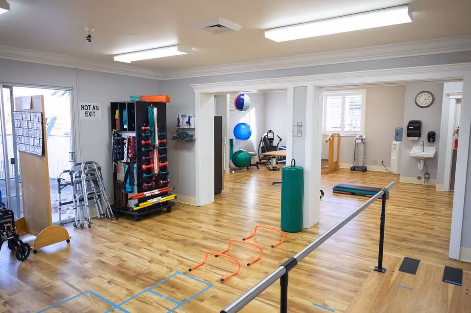 A spacious rehabilitation or physical therapy room with wooden floors, exercise equipment including dumbbells, resistance bands, exercise balls, and walkers. There is a parallel bar for walking exercises, a sink with soap dispensers, a clock on the wall, and a sign that reads 'NOT AN EXIT' near a glass door.