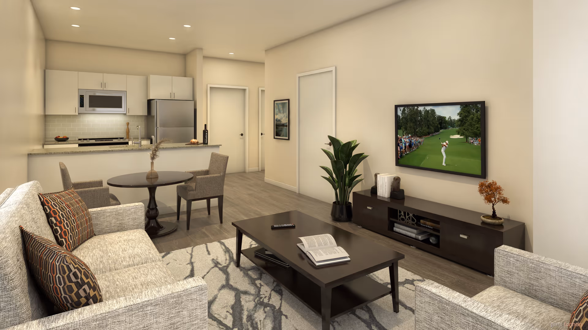 A modern senior living apartment interior featuring a living room with a gray sofa and armchair, a dark wood coffee table with an open book and remote control, a flat-screen TV mounted on the wall showing a golf game, and a dark wood TV console with books and a small plant. In the background, there is a small round dining table with two chairs and a kitchen area with white cabinets, a microwave, and a stainless steel refrigerator. The room has light-colored walls and wood flooring.