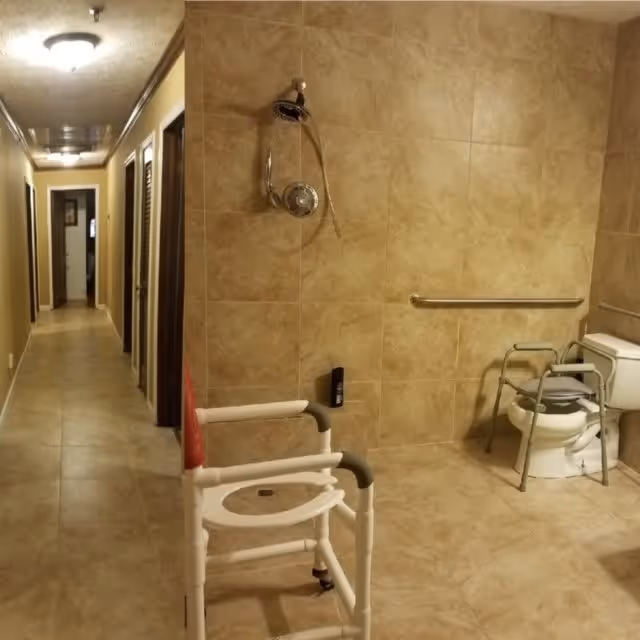 A spacious bathroom with beige tiled walls and floor, featuring a showerhead, a white shower chair, a toilet with a metal support frame, and a horizontal grab bar on the wall. To the left is a hallway with multiple doors and ceiling lights.