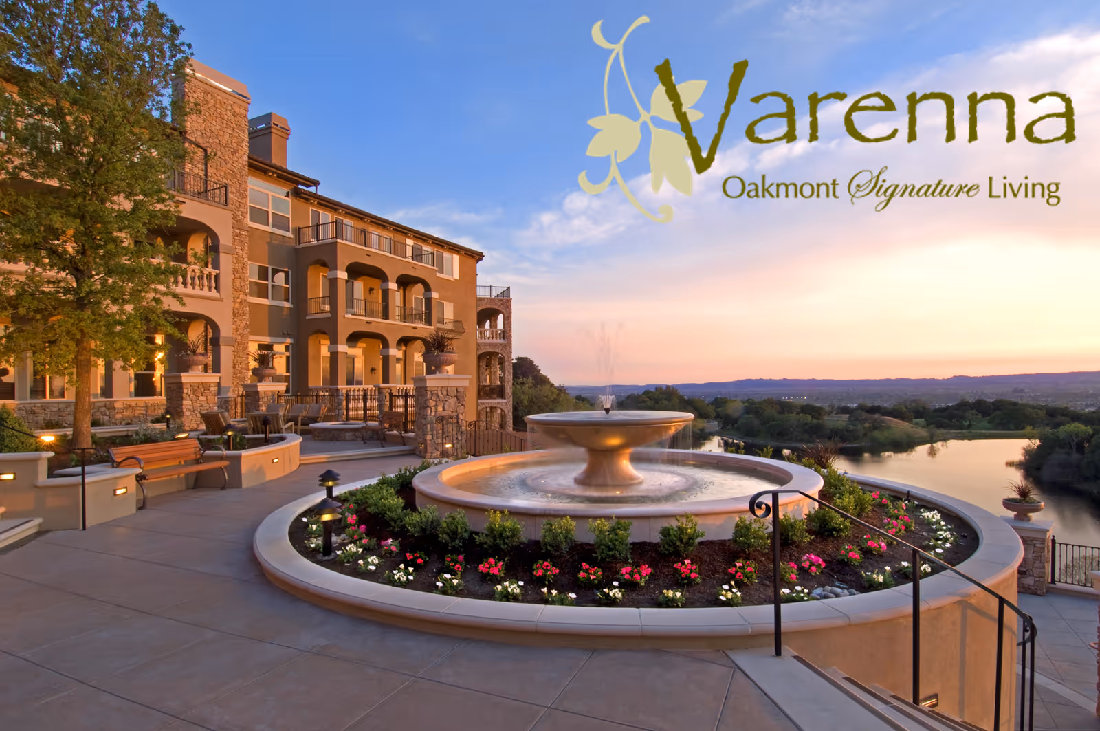 Outdoor view of a senior living facility named Varenna at Fountaingrove during sunset, featuring a large circular fountain surrounded by flowers and greenery, with a multi-story building and a scenic river and hills in the background.