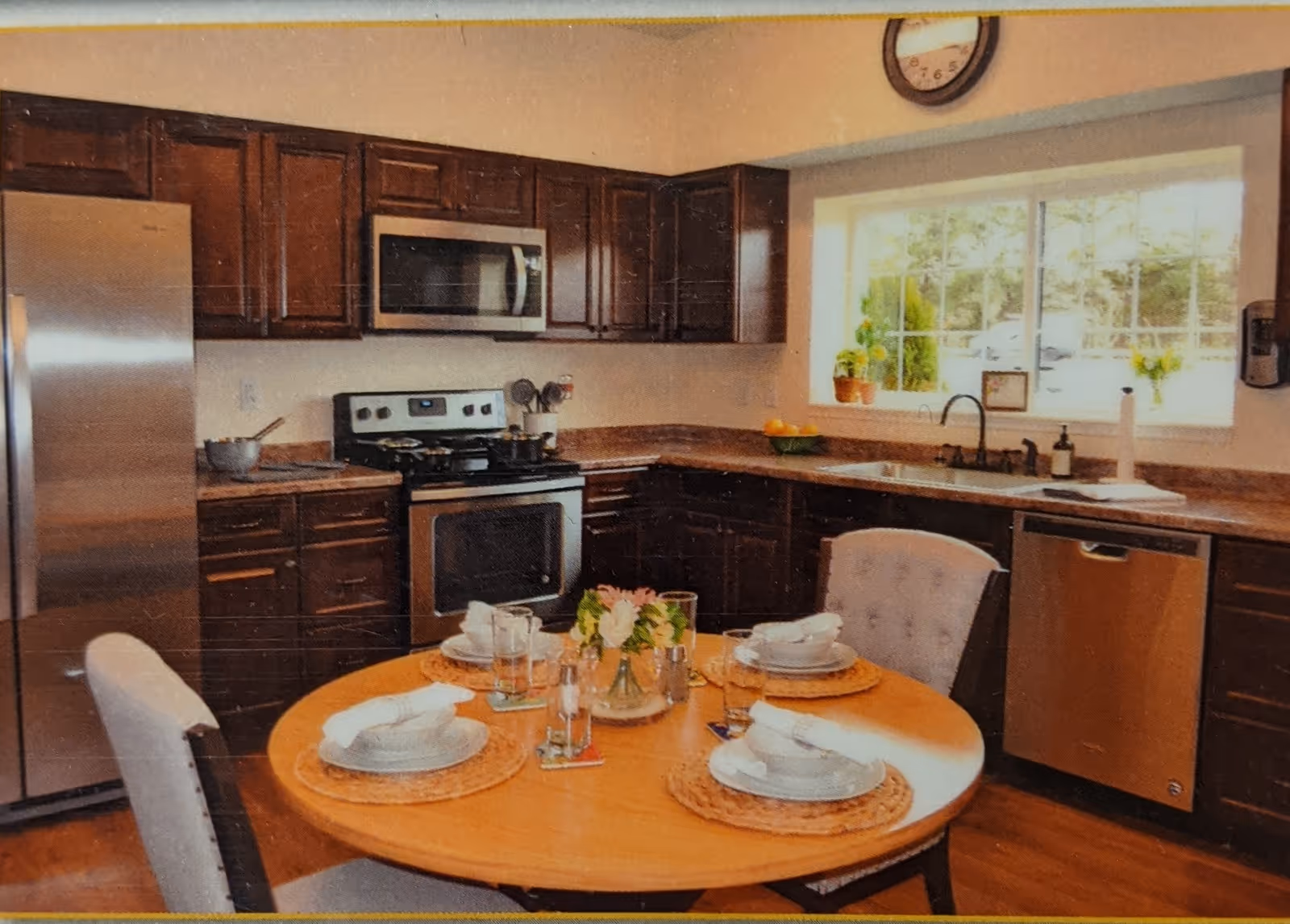 Well-lit kitchen with stainless steel appliances, dark cabinets, a round dining table set for four, and a window above the sink.