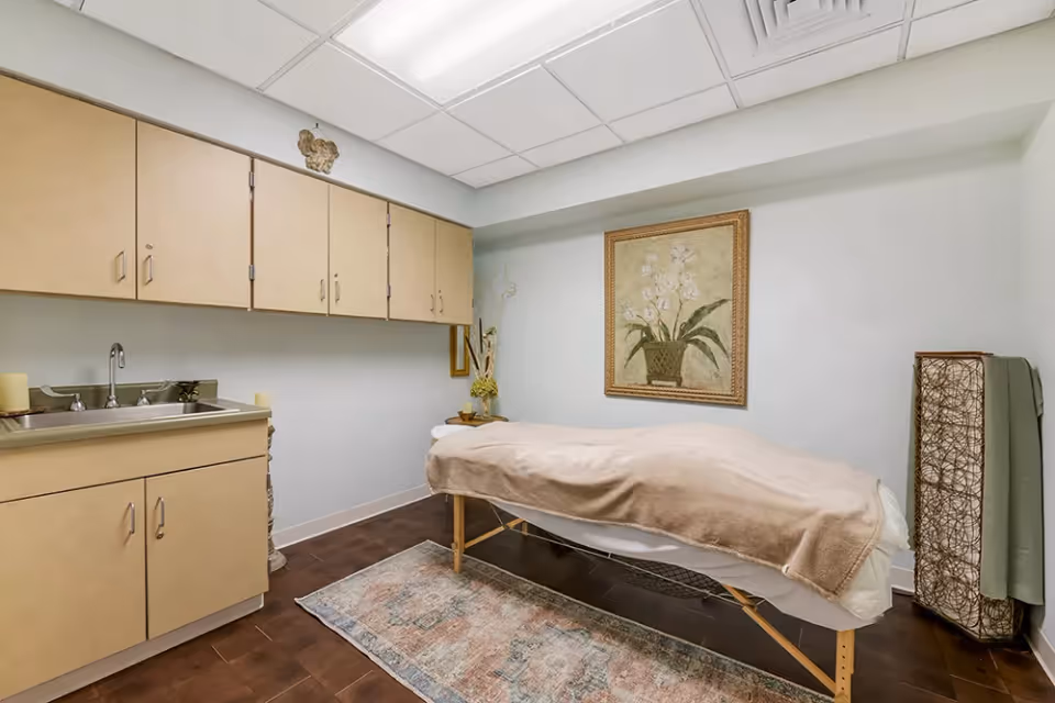 A small room with a massage or treatment table covered with a beige blanket, a framed floral painting on the wall, beige cabinets with a sink on the left side, and a decorative lamp with a green cloth on the right side. The floor has a patterned rug and the ceiling has white tiles with a vent.